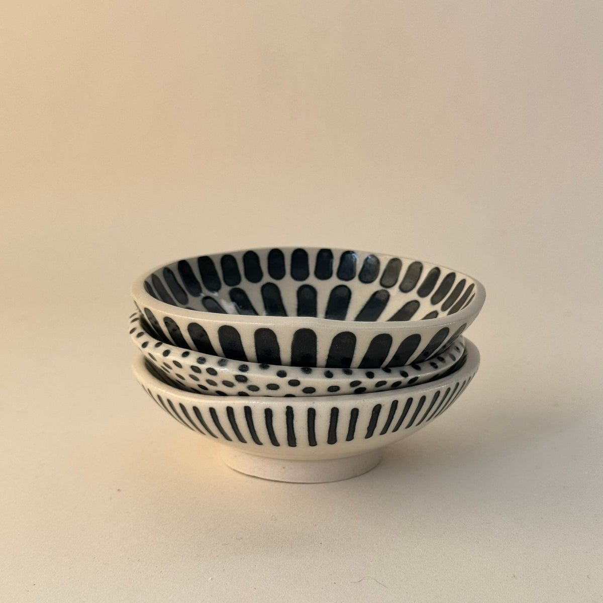 Tre staplade handgjorda skålar i stengods med handmålade svarta prickar, breda och smala ränder samt transparent glasyr, fotograferade snett framifrån.