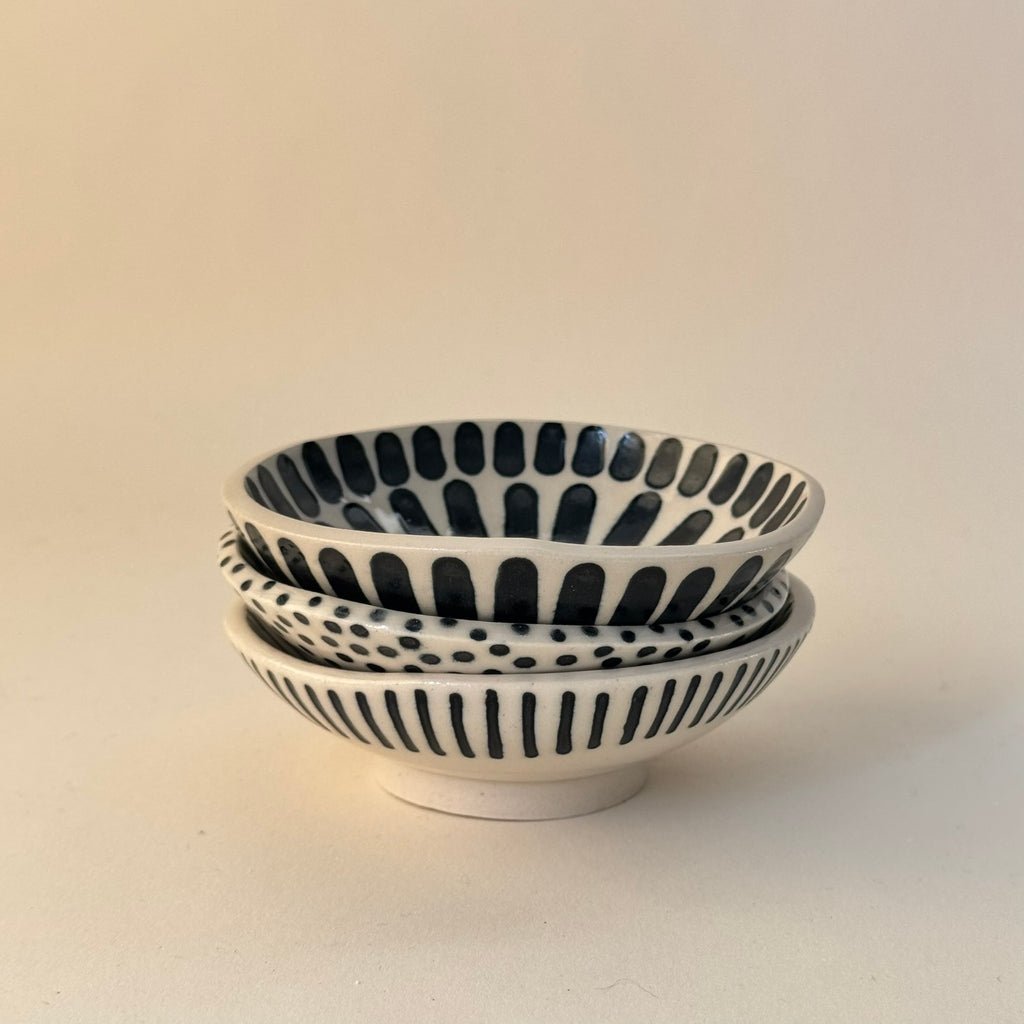 Tre staplade handgjorda skålar i stengods med handmålade svarta prickar, breda och smala ränder samt transparent glasyr, fotograferade snett framifrån.