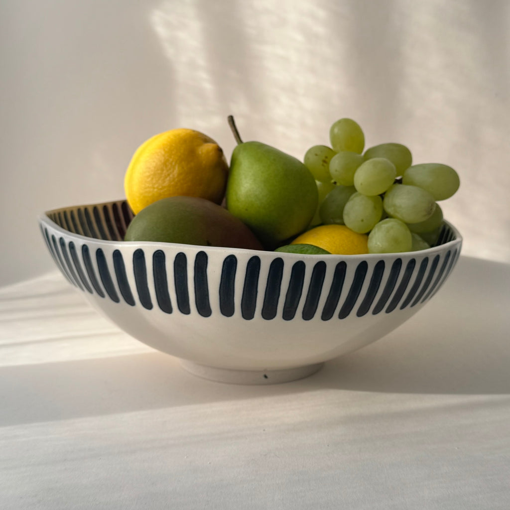 Handgjord skål i stengods med breda handmålade svarta ränder och transparent glasyr, fylld med färsk frukt och gröna vindruvor.