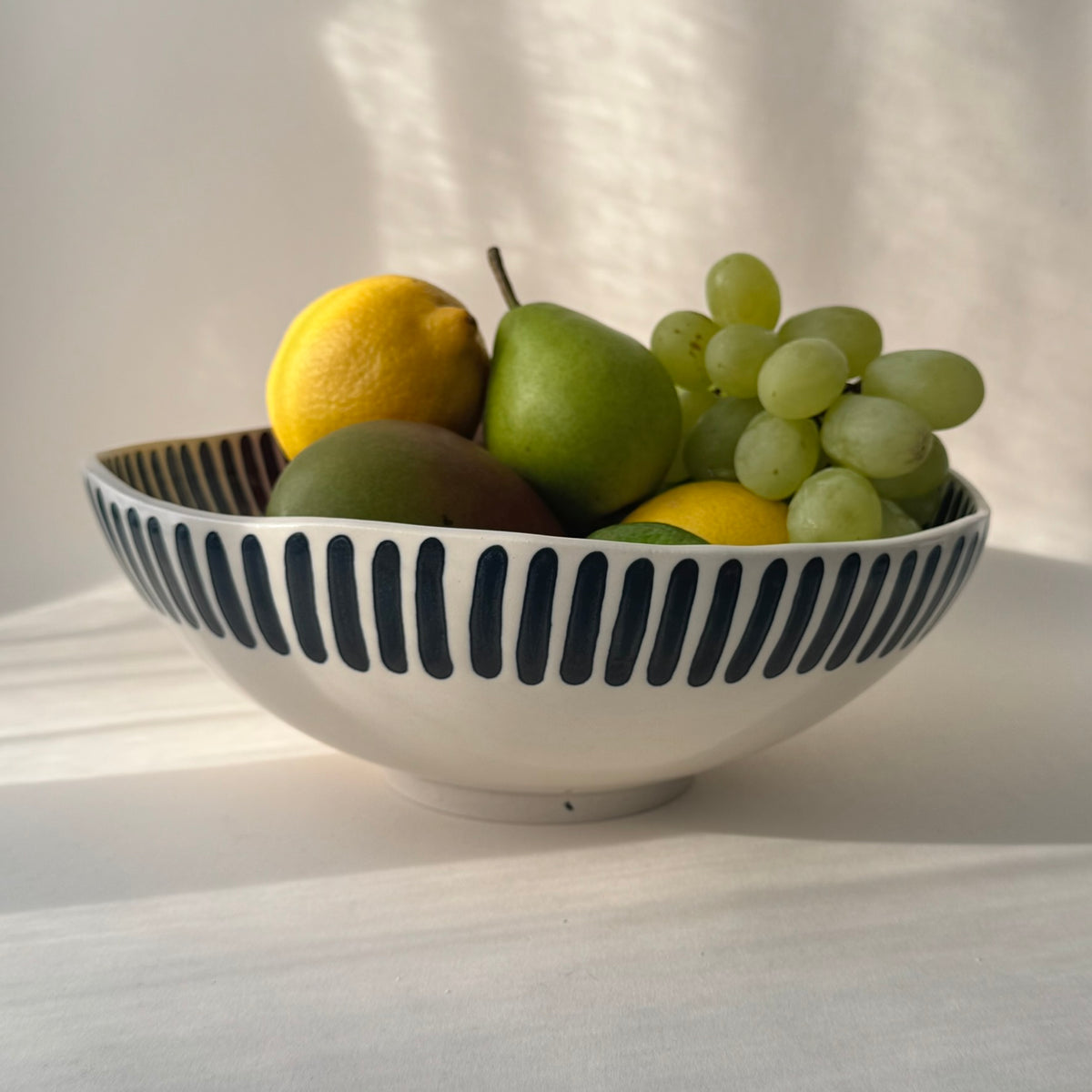 Handgjord skål i stengods med breda handmålade svarta ränder och transparent glasyr, fylld med färsk frukt och gröna vindruvor.