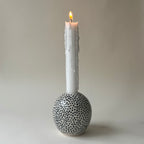 Handgjord rund ljusstake i stengods med handmålade svarta prickar och transparent glasyr, med ett tänt vitt kronljus.